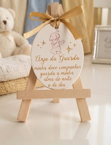 lembrança oração santo anjo com mini cavalete encante seus convidados com esta delicada e significativa lembrança, perfeita para batizados, nascimentos e primeiras comunhões. detalhes do produto: design exclusivo: uma placa oval em acabamento branco acetinado, apresentando uma ilustração de um anjo orando em relevo e detalhes em brilho rose gold. oração gravada: inclui a bela e clássica oração em português: "anjo da guarda, minha doce companhia, guarda a minha alma de noite e de dia!" toque final: um sofisticado laço de fita de cetim dourado na parte superior confere charme extra à peça. acompanhamento: acompanha um mini cavalete de madeira natural, ideal para expor a lembrança com elegância em qualquer ambiente. apresentação profissional: exibida em um ambiente de berçário acolhedor e fofo, como mantas suaves, um ursinho de pelúcia e um porta retratos, demonstrando como ela ficará perfeita na decoração. ideal para: lembrancinhas de batizado lembrancinhas de nascimento presentes de primeira comunhão decoração de quartos infantis traga um toque de espiritualidade, proteção e beleza para celebrar momentos especiais com esta lembrança única.